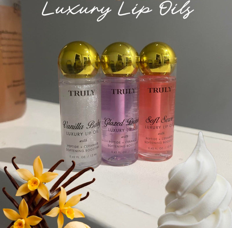 Three bottles of Truly luxury lip oils with gold caps on a white surface with decorative elements.
